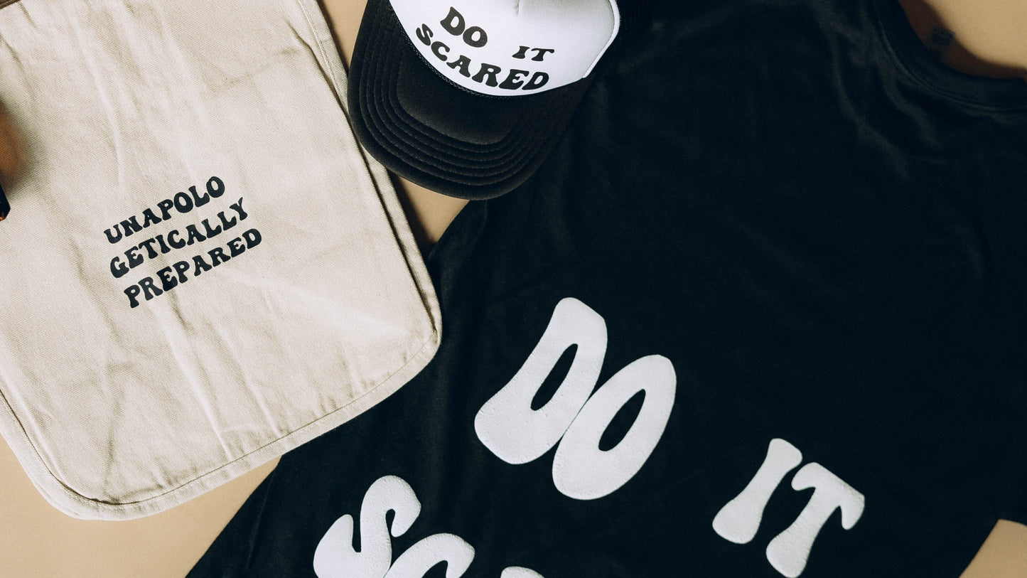 Black t-shirt with 'DO IT SCARED' text, beige tote bag, and black sneakers on a beige surface.