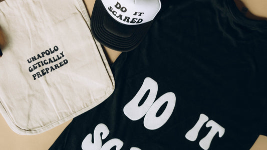 Black t-shirt with 'DO IT SCARED' text, beige tote bag, and black sneakers on a beige surface.